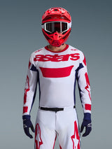 Motocross rider wearing the Alpinestars 2026 Racer Riway Jersey in White Blue Red, featuring a large red 'astars' logo on the chest, navy blue side panels, matching white and red motocross pants, a red helmet with white goggles, and navy blue gloves, complete off-road racing gear set.