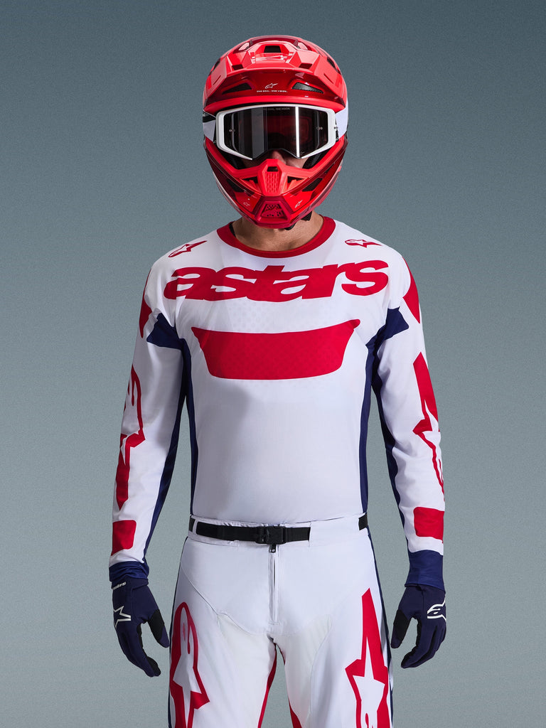 Pilota di motocross che indossa la maglia Alpinestars 2026 Racer Riway in White Blue Red, caratterizzata da un grande logo 'astars' rosso sul petto, pannelli laterali blu navy, pantaloni da motocross coordinati bianchi e rossi, casco rosso con occhiali bianchi e guanti blu navy, set completo da gara off-road.