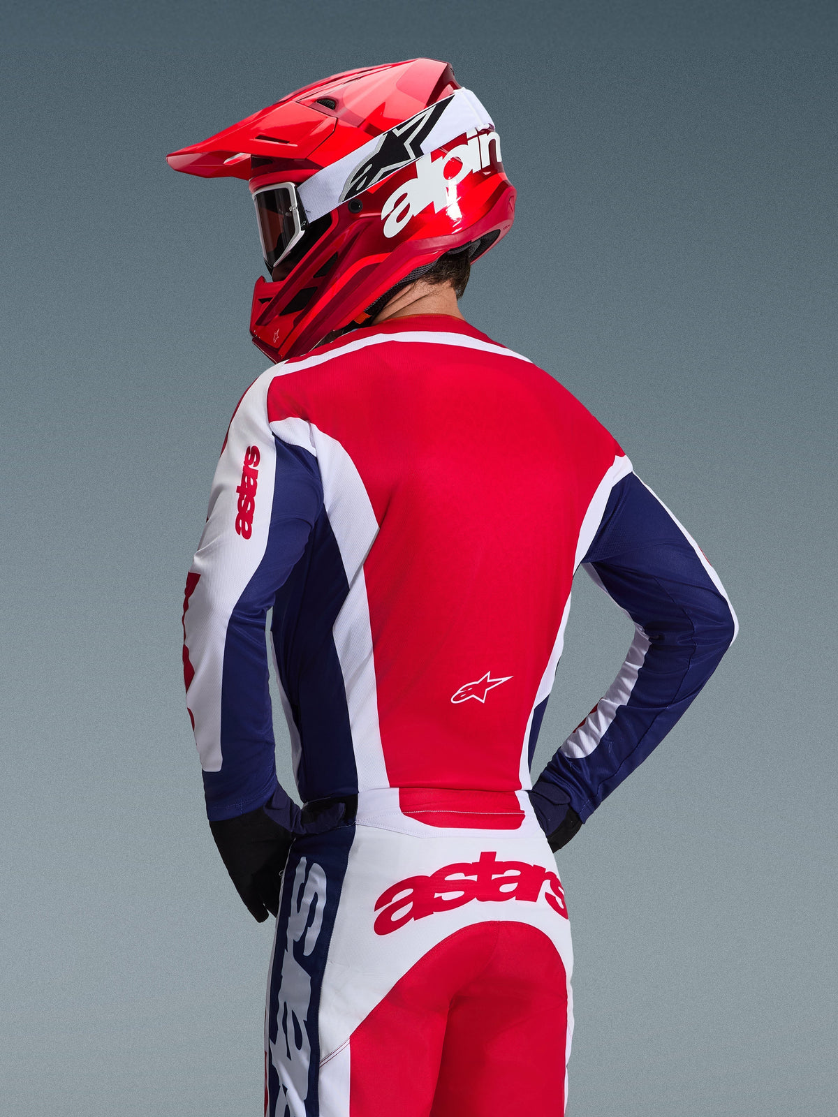 Pilota di motocross visto da dietro con maglia Alpinestars 2026 Racer Riway, colorazione bianco, blu e rosso, pantaloni da fuoristrada coordinati con logo astars, casco rosso con logo Alpinestars bianco, guanti neri, equipaggiamento da motocross coordinato