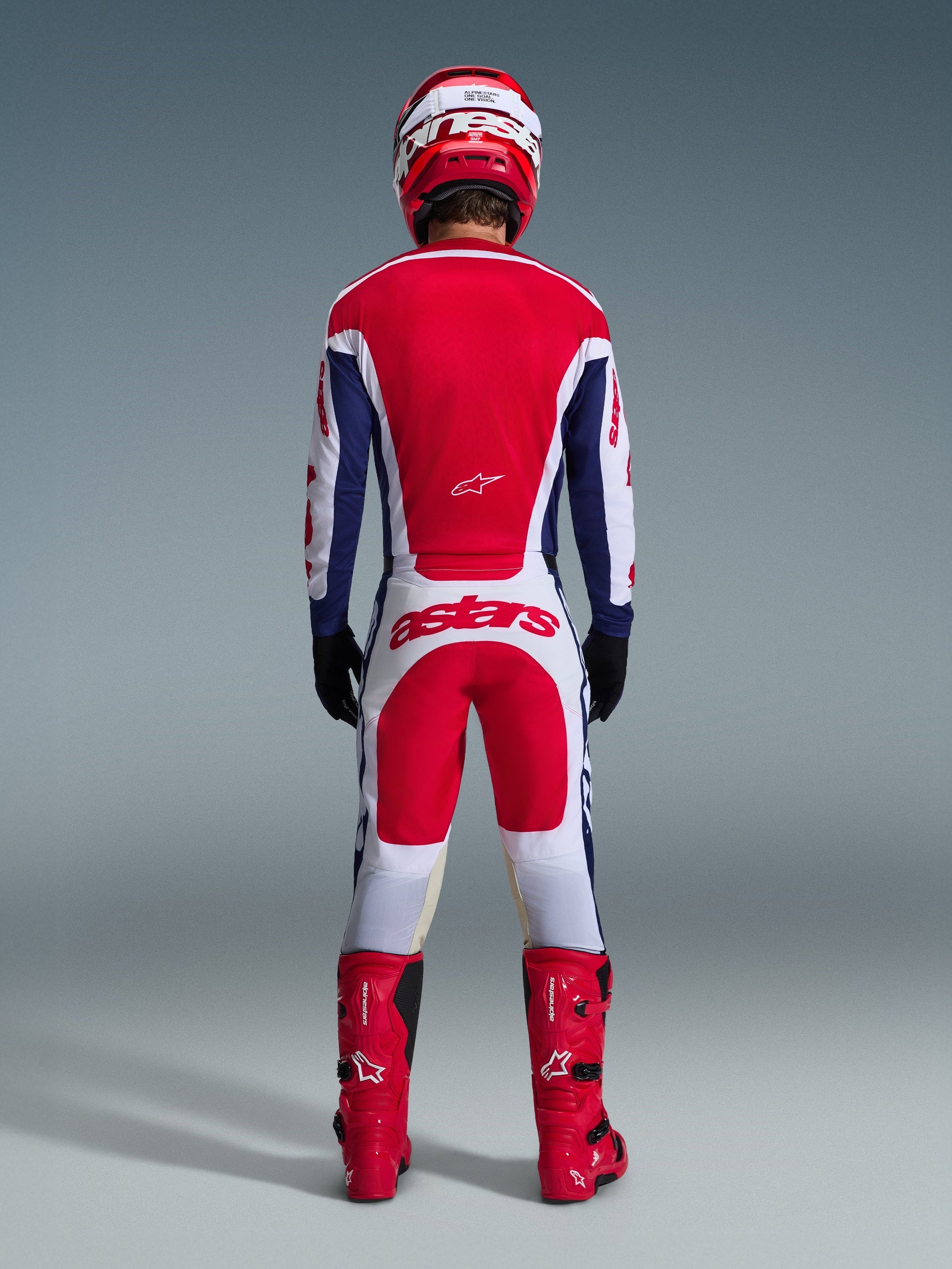 Motocross rider wearing complete racing gear from behind, Alpinestars 2026 Racer Riway Jersey in White Blue Red, matching white and red pants with bold 'astars' branding, red boots, and red and white helmet, coordinated off-road racing outfit.