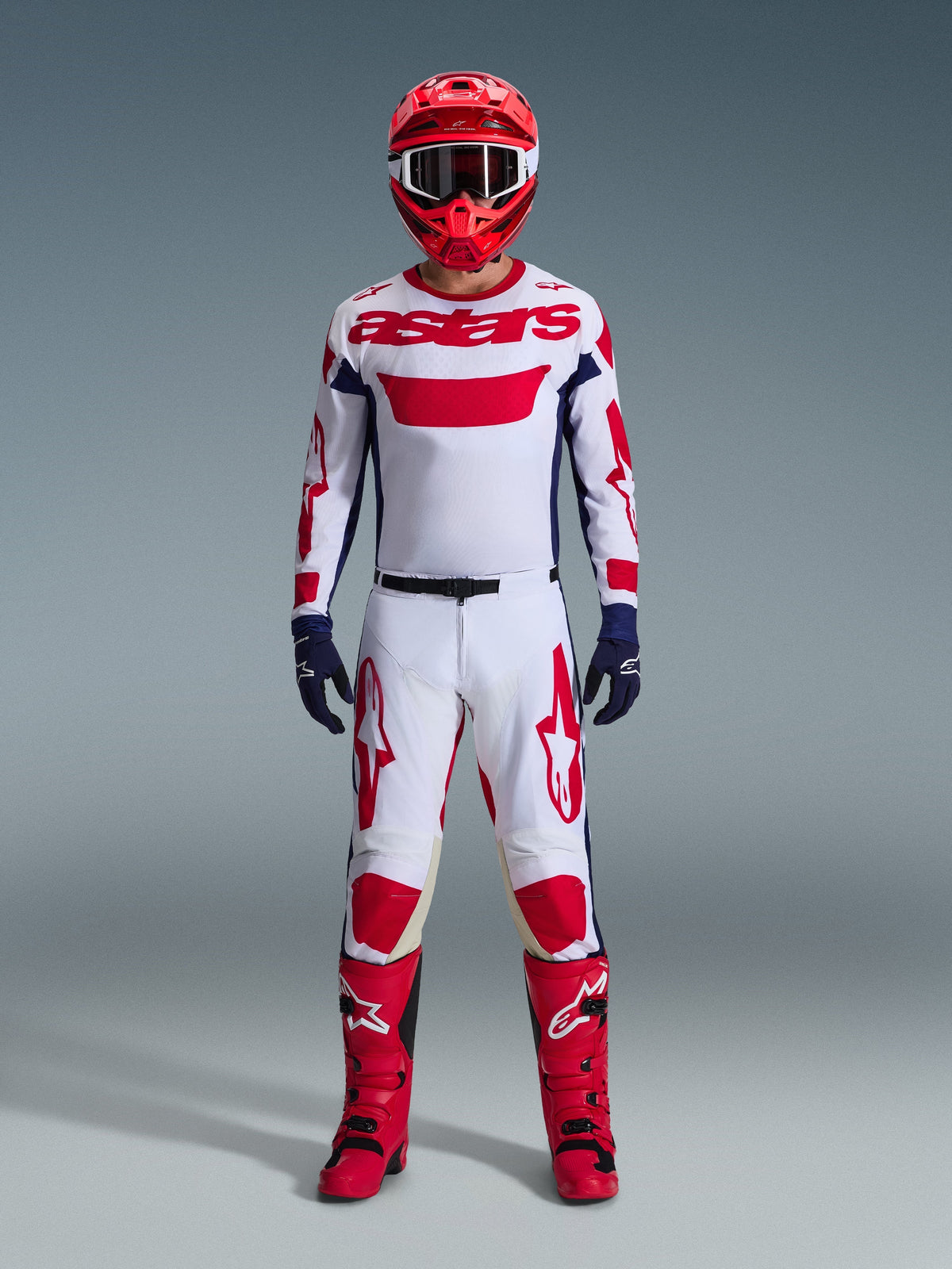 Pilota di motocross con abbigliamento completo da gara, maglia Alpinestars 2026 Racer Riway in Bianco Blu Rosso, pantaloni bianchi e rossi abbinati, casco rosso, stivali rossi e guanti blu navy, completo coordinato per corse fuoristrada