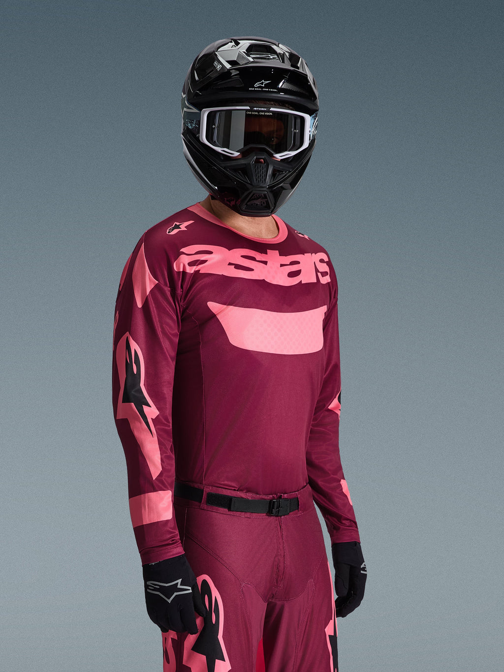 Pilota di motocross che indossa il completo Alpinestars Racer Riway 2026, maglia bordeaux e rosa con logo 'astars' in evidenza, pantaloni coordinati con dettagli rosa, casco e guanti neri, abbigliamento da gara professionale per motocross off-road