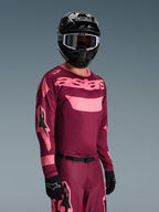Pilota di motocross che indossa il completo Alpinestars Racer Riway 2026, maglia bordeaux e rosa con logo 'astars' in evidenza, pantaloni coordinati con dettagli rosa, casco e guanti neri, abbigliamento da gara professionale per motocross off-road