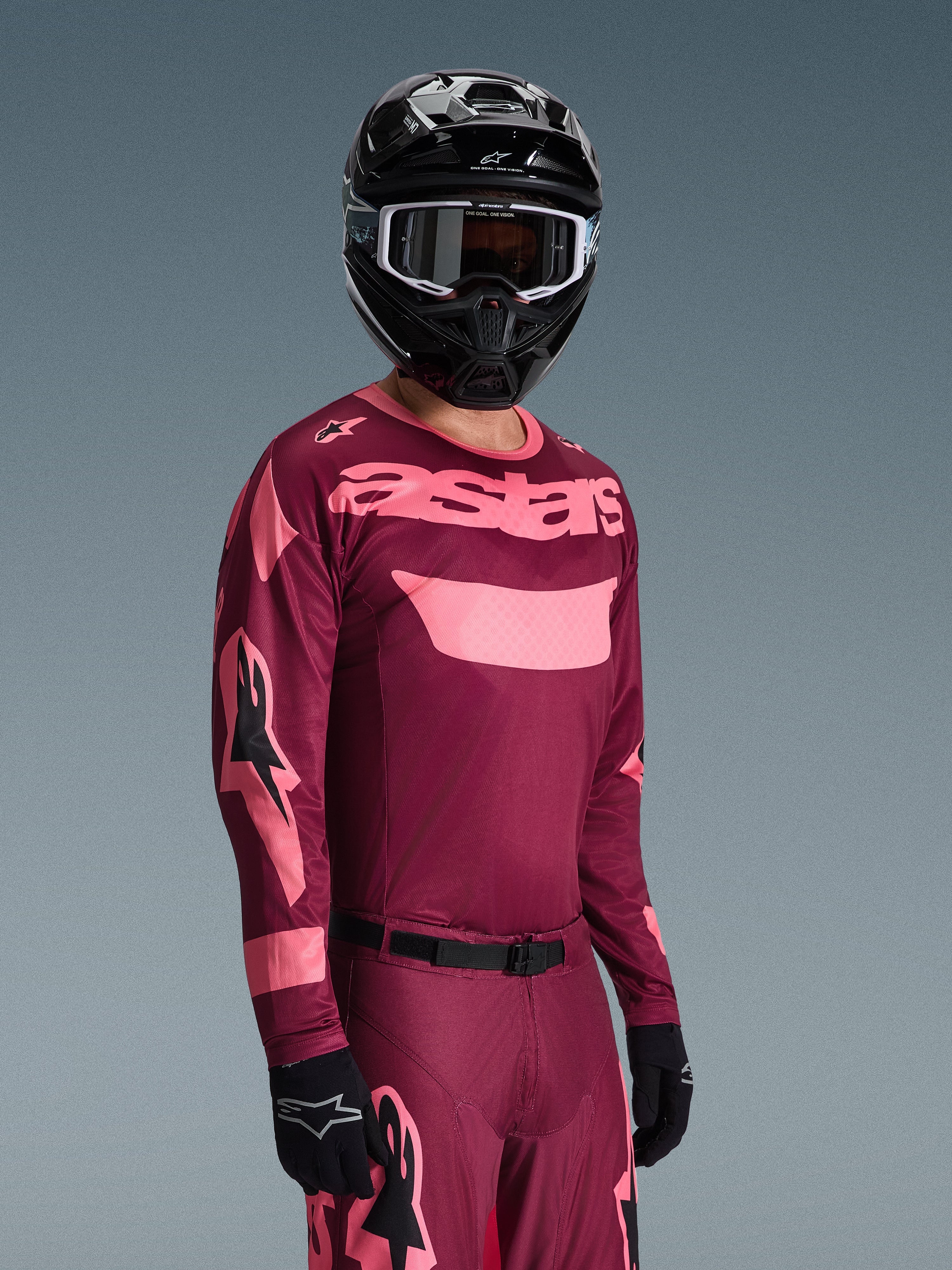 Pilota di motocross che indossa il completo Alpinestars Racer Riway 2026, maglia bordeaux e rosa con logo 'astars' in evidenza, pantaloni coordinati con dettagli rosa, casco e guanti neri, abbigliamento da gara professionale per motocross off-road