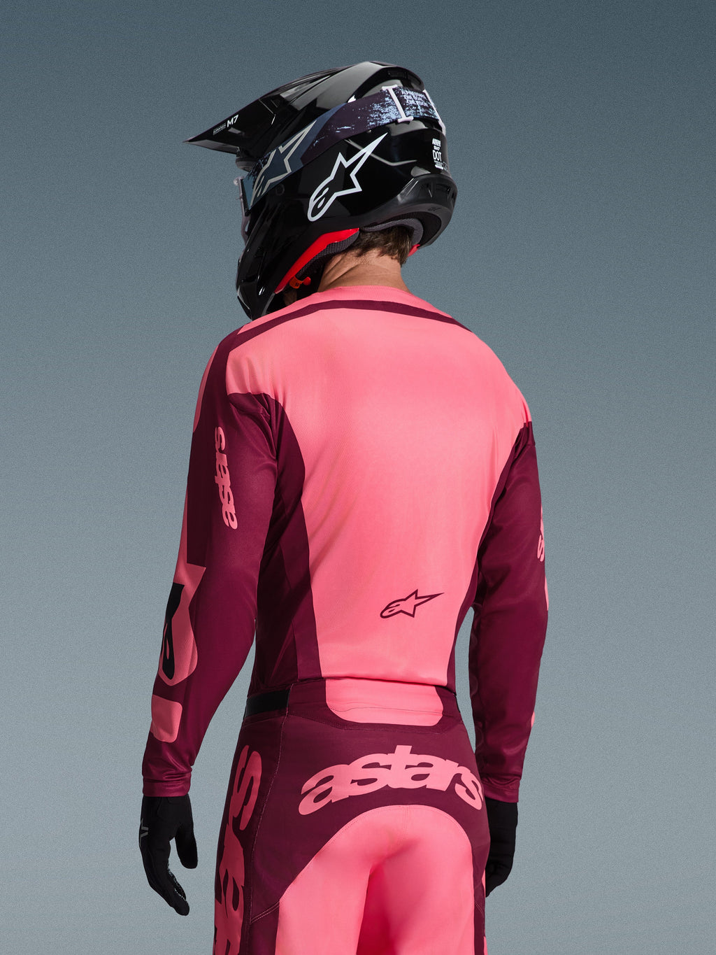 Pilota di motocross visto da dietro con il completo Alpinestars 2026 Racer Riway, maglia e pantaloni rosa acceso e bordeaux, casco da motocross nero con logo bianco e guanti neri, con marchio 'astars' sulla schiena.