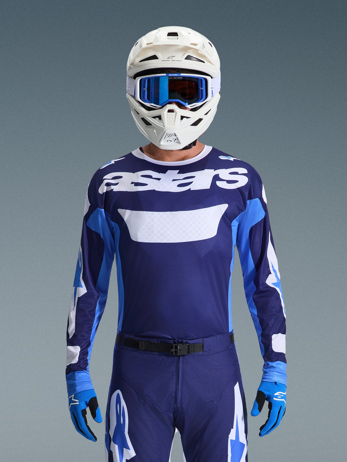 Pilota di motocross con maglia Alpinestars Racer Riway 2026, blu UCLA e bianco con logo 'astars' bianco sul petto, casco bianco con occhiali blu, pantaloni da motocross e guanti blu coordinati, abbigliamento da gara off-road completo