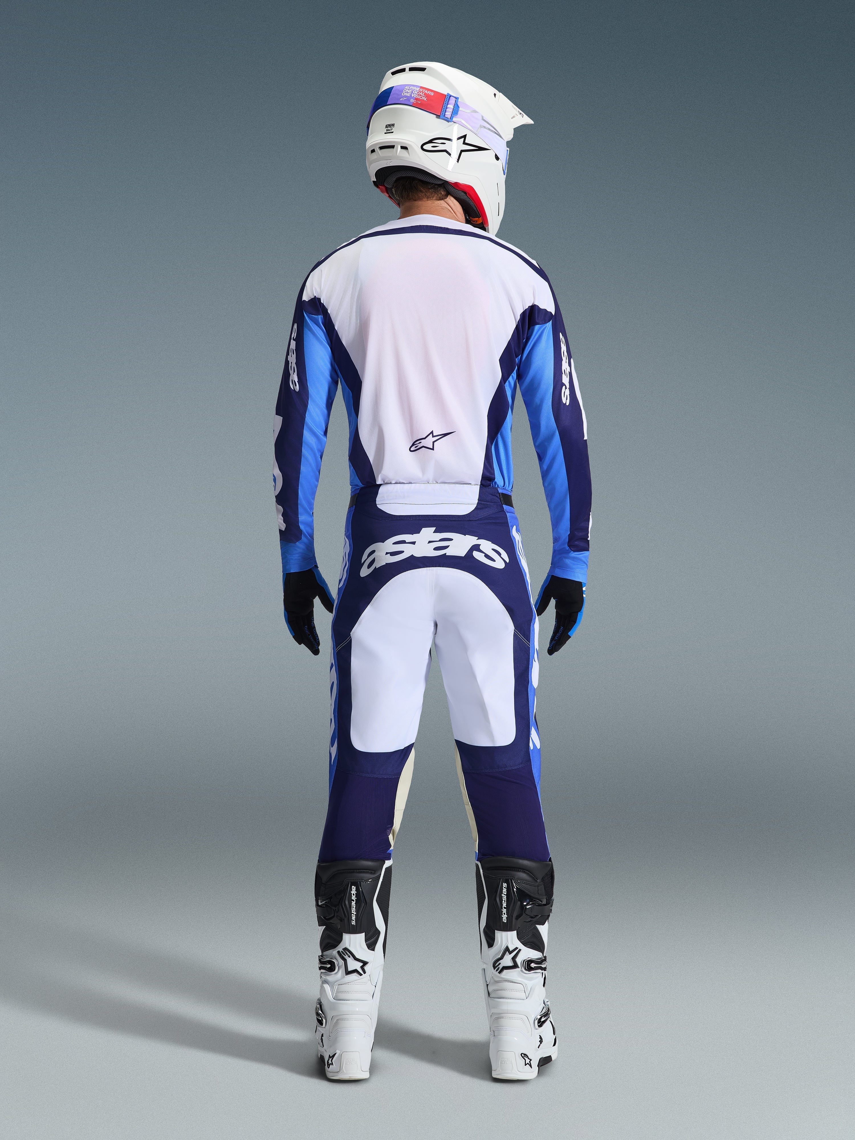 Pilota di motocross visto da dietro che indossa la maglia Alpinestars 2026 Racer Riway in Ucla Blue e bianco, pantaloni da motocross coordinati con pannelli bianchi e blu navy e marchio Astars, stivali bianchi e neri e casco bianco, completo da gara coordinato per prestazioni off-road.