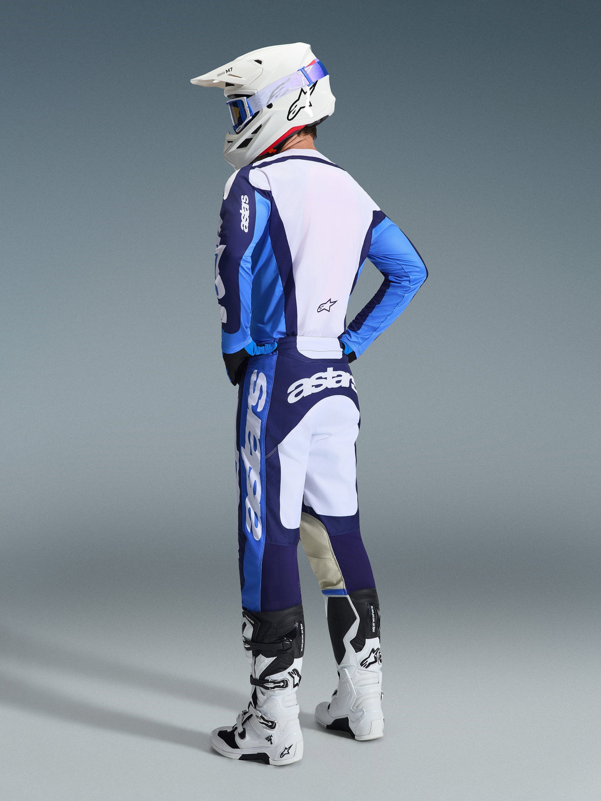 Pilota di motocross che indossa la maglia Racer Riway 2026 in blu Ucla e bianco, abbinata a pantaloni da motocross coordinati blu e bianchi, un casco bianco e stivali bianchi, mostrato da una vista posteriore di tre quarti per un look completo da gara off-road.