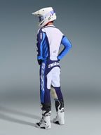 Pilota di motocross che indossa la maglia Racer Riway 2026 in blu Ucla e bianco, abbinata a pantaloni da motocross coordinati blu e bianchi, un casco bianco e stivali bianchi, mostrato da una vista posteriore di tre quarti per un look completo da gara off-road.