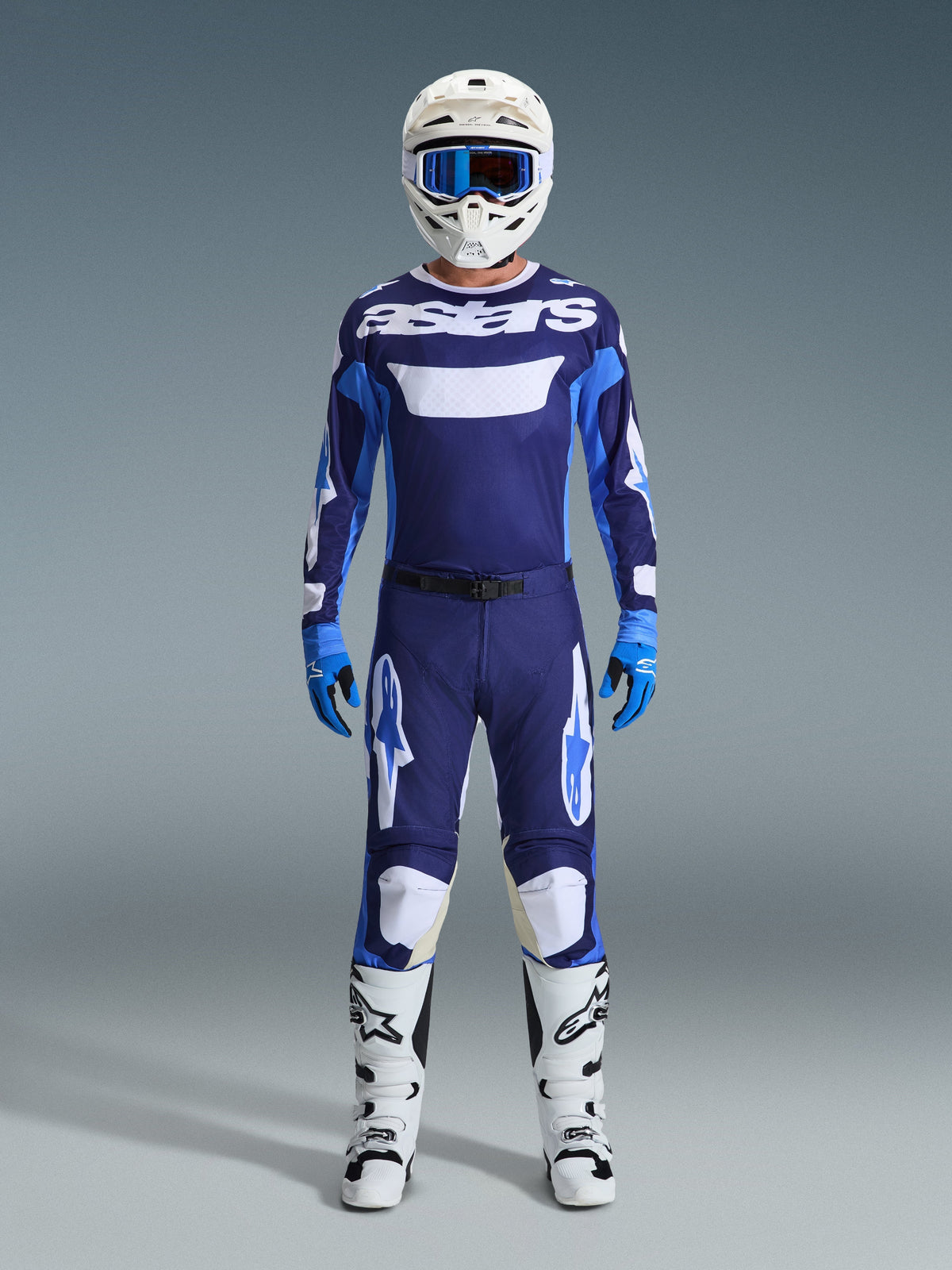 Pilota di motocross con abbigliamento completo da gara, Maglia Racer Riway 2026 in blu UCLA e bianco con inserti navy e branding 'astars' sul petto, pantaloni coordinati blu e bianchi, casco bianco con maschera blu, stivali bianchi e neri, guanti blu, kit da motocross coordinato.