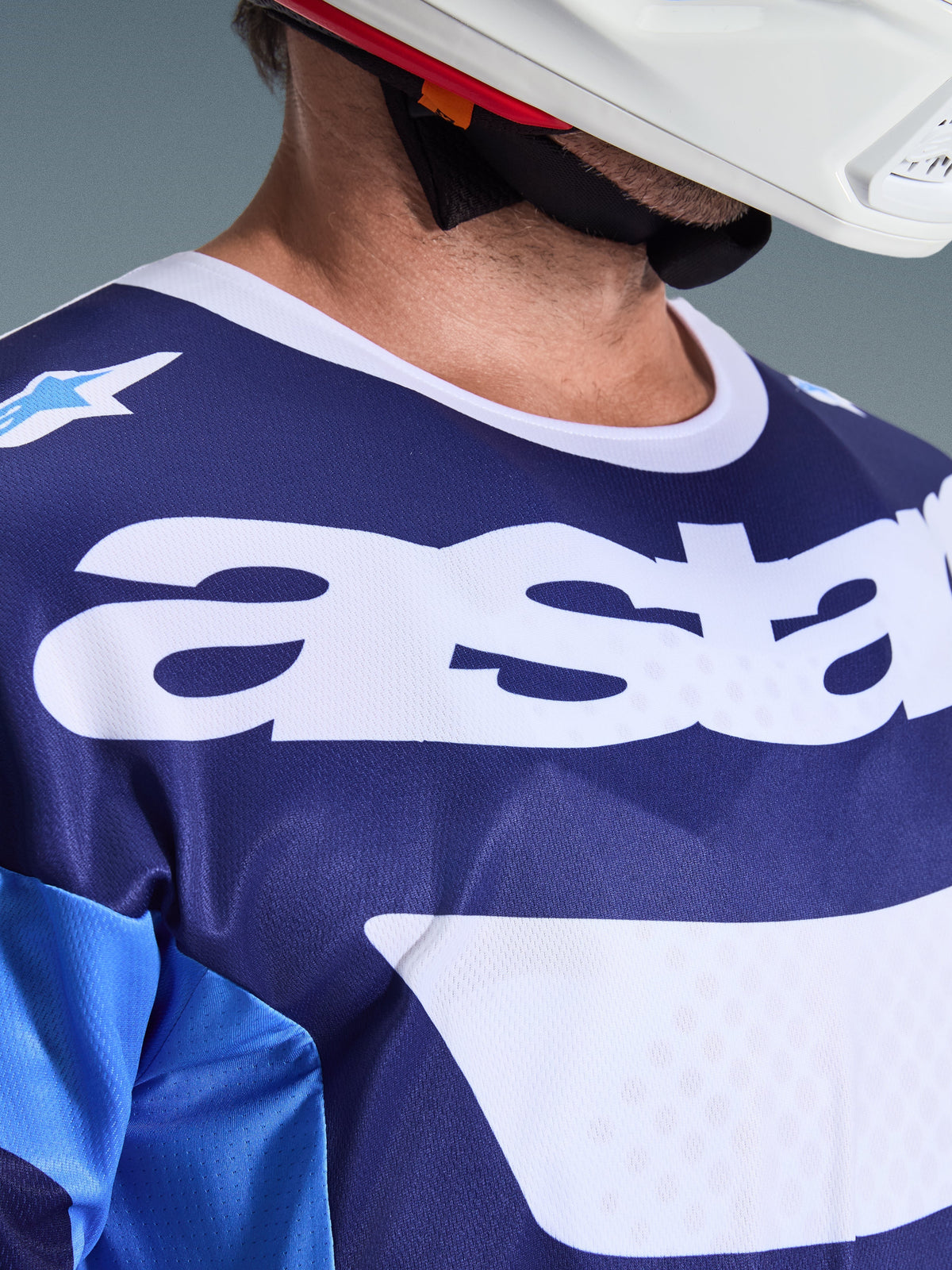 Primo piano della maglia Alpinestars Racer Riway 2026 in blu UCLA e bianco, con grande logo 'astars' bianco sul petto, colletto bianco e trama in rete traspirante, indossata con un casco da motocross bianco.