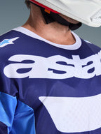 Primo piano della maglia Alpinestars Racer Riway 2026 in blu UCLA e bianco, con grande logo 'astars' bianco sul petto, colletto bianco e trama in rete traspirante, indossata con un casco da motocross bianco.