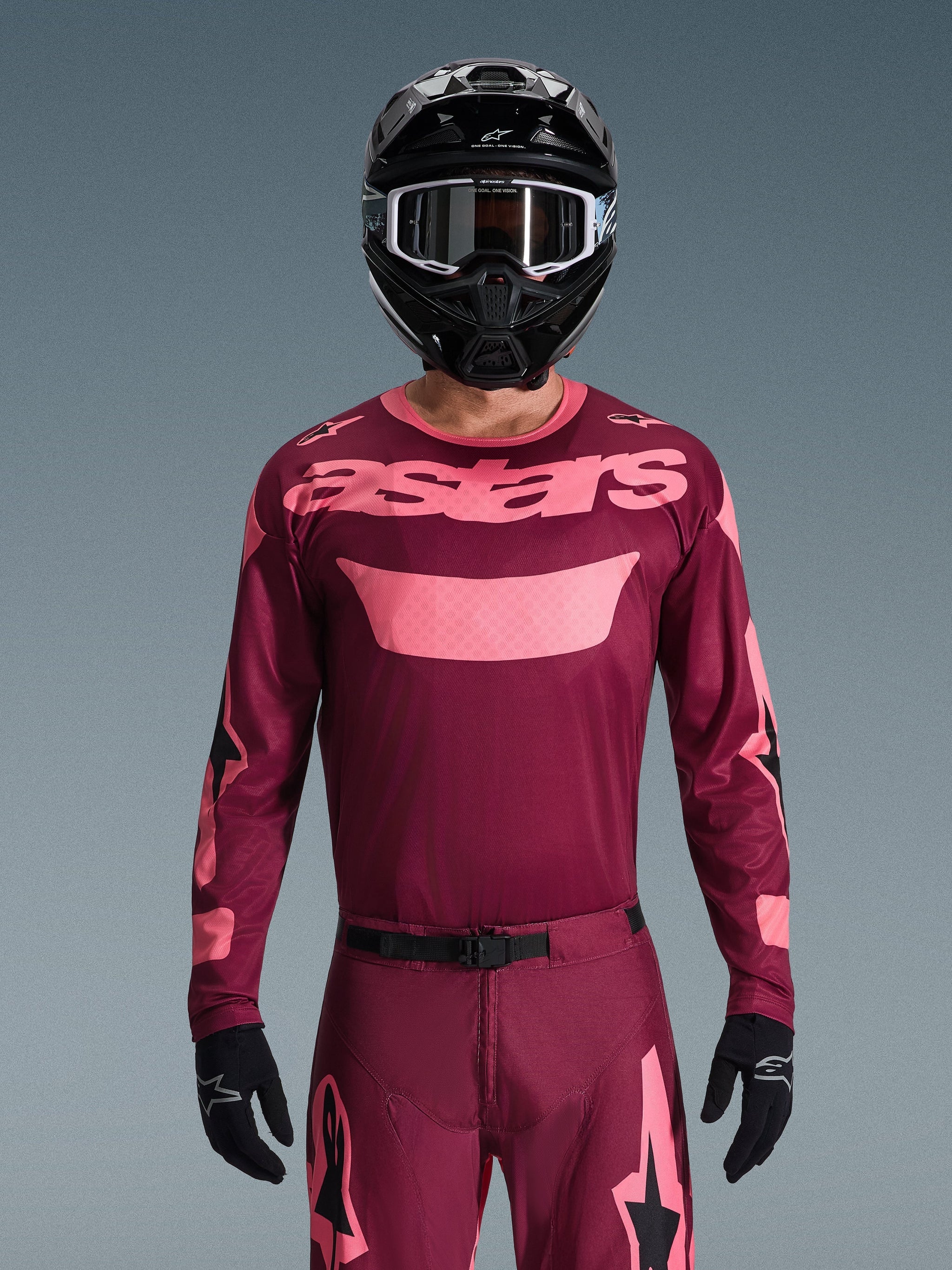 Pilota di motocross che indossa la maglia Alpinestars Racer Riway 2026 in Dark Red Coral, con corpo bordeaux e logo 'astars' rosa corallo sul petto, abbinata a pantaloni coordinati, casco nero e guanti neri, set professionale per fuoristrada.