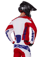 Maglia e pantaloni Alpinestars Racer Veil 2025, completo da motocross, rosso acceso, bianco e blu, vista posteriore del pilota che indossa il completo coordinato e un casco da motocross nero, con branding Alpinestars blu in evidenza, abbigliamento tecnico da corsa off-road