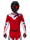 Maglia Alpinestars Racer Veil 2025, maglia da motocross, rossa, bianca e blu, a maniche lunghe, indossata con casco nero e maschera arancione, guanti e pantaloni rossi coordinati, design sportivo ad alte prestazioni per la guida fuoristrada