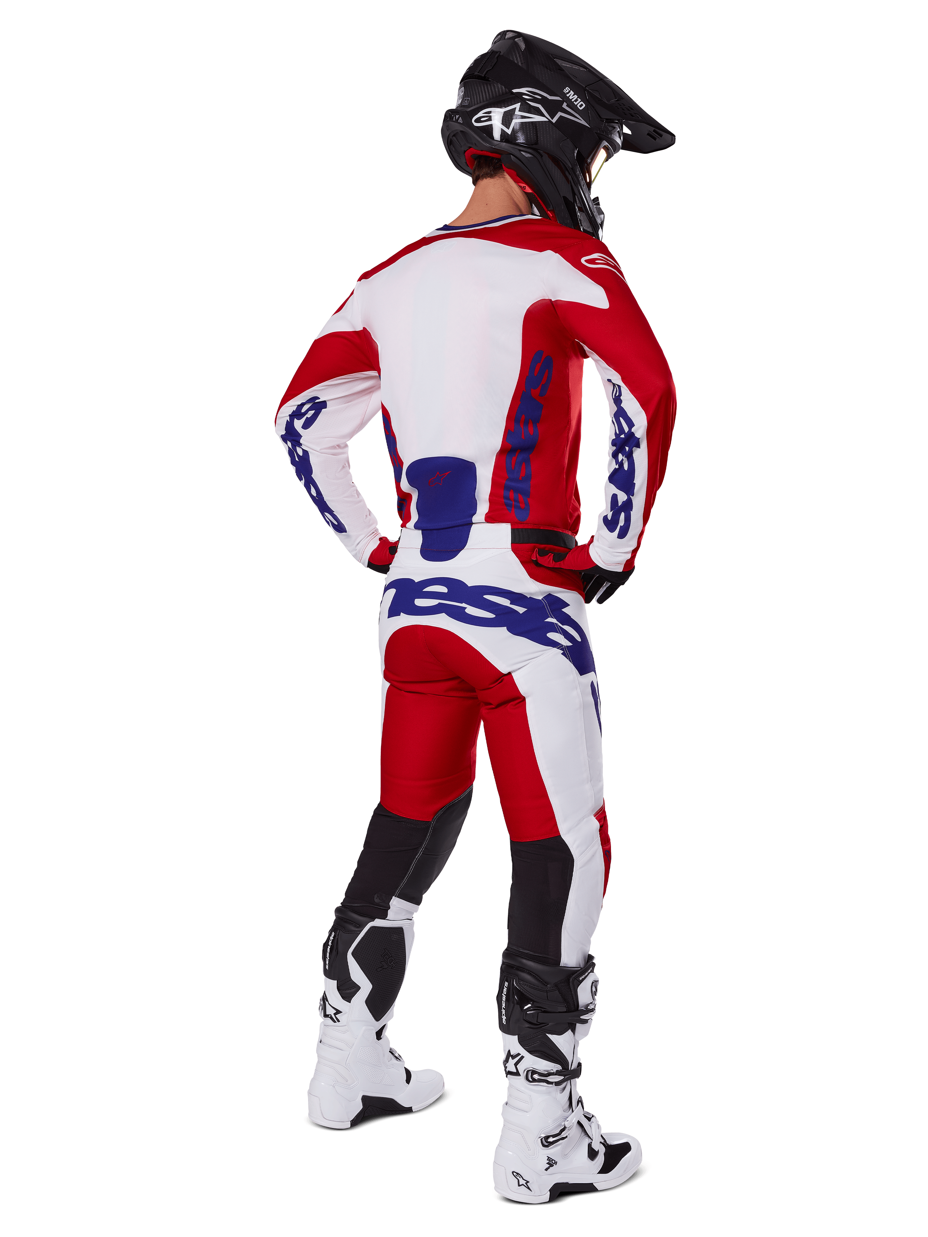 Pilota di motocross visto da dietro che indossa la maglia e i pantaloni Alpinestars Racer Veil 2025 in rosso e bianco, con loghi blu, casco nero e stivali bianchi, set completo di abbigliamento per corse fuoristrada