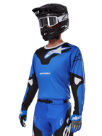 Alpinestars 2025 Racer Veil Gear Set, maglia da motocross blu con grafiche bianche e nere, indossata con pantaloni coordinati, casco nero, occhiali bianchi e guanti blu, abbigliamento tecnico per corse fuoristrada