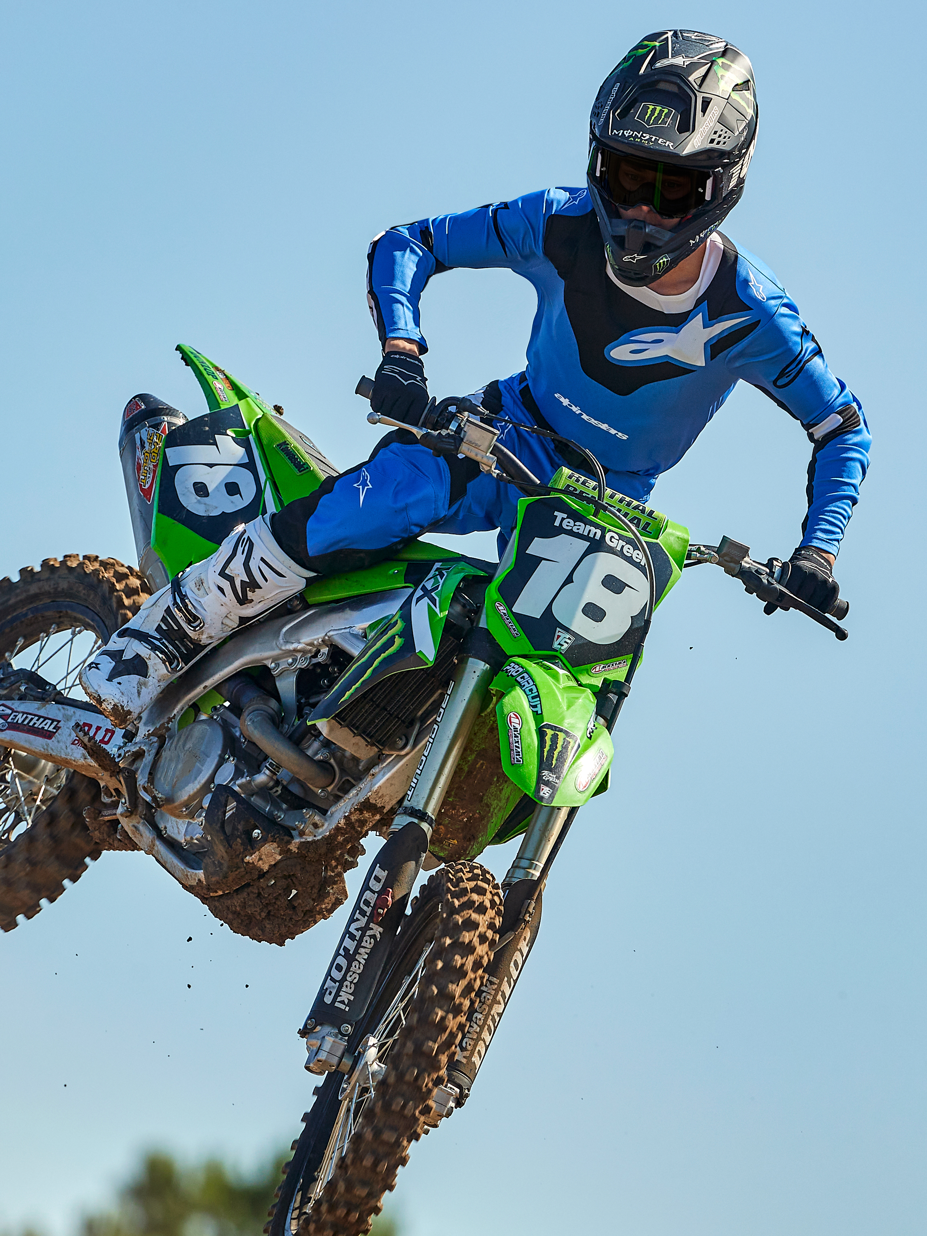 Alpinestars 2025 Racer Veil Jersey, maglia da motocross, blu e nera, maglia tecnica a maniche lunghe con grafiche astratte e sezioni in rete traspirante, indossata da un pilota durante un salto off-road su una moto verde