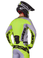 Pilota di motocross visto da dietro che indossa la maglia Alpinestars 2025 Racer Veil in grigio e giallo fluo, con l'evidente marchio 'astars' sulla schiena e sulle maniche, abbinata a pantaloni coordinati e un casco nero per un look racing completo.