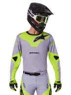 Alpinestars Racer Veil Jersey 2025, maglia da motocross, grigio e giallo neon, design a maniche lunghe con loghi Alpinestars neri, mostrata come parte di un set completo da gara con pantaloni coordinati, casco nero e guanti.