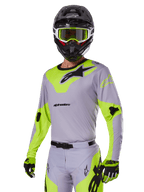 Pilota di motocross con set completo Alpinestars Racer Veil 2025 grigio e verde fluo, maglia e pantaloni grigio chiaro con pannelli giallo-verde neon e loghi neri, guanti abbinati, casco nero con occhiali verde neon, abbigliamento professionale da gara fuoristrada