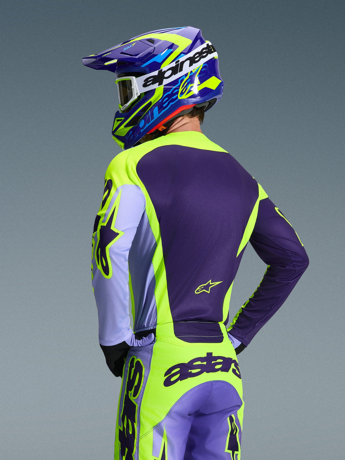 Pilota di motocross visto da dietro con maglia Alpinestars 2026 Racer Portl e pantaloni coordinati, colorazione viola e giallo fluo con accenti lavanda, casco coordinato blu e giallo, abbigliamento race-fit per prestazioni off-road.