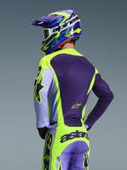 Pilota di motocross visto da dietro con maglia Alpinestars 2026 Racer Portl e pantaloni coordinati, colorazione viola e giallo fluo con accenti lavanda, casco coordinato blu e giallo, abbigliamento race-fit per prestazioni off-road.