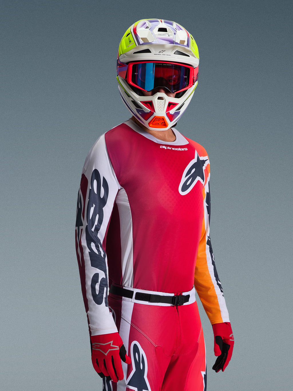 Pilota di motocross che indossa il completo Alpinestars 2026 Racer Portl, maglia rossa con sfumature rosa e maniche bianche, pantaloni rossi coordinati, casco bianco con vivaci accenti gialli e viola, occhiali rossi con lenti blu iridescenti e guanti rossi, abbigliamento da gara professionale per motocross.