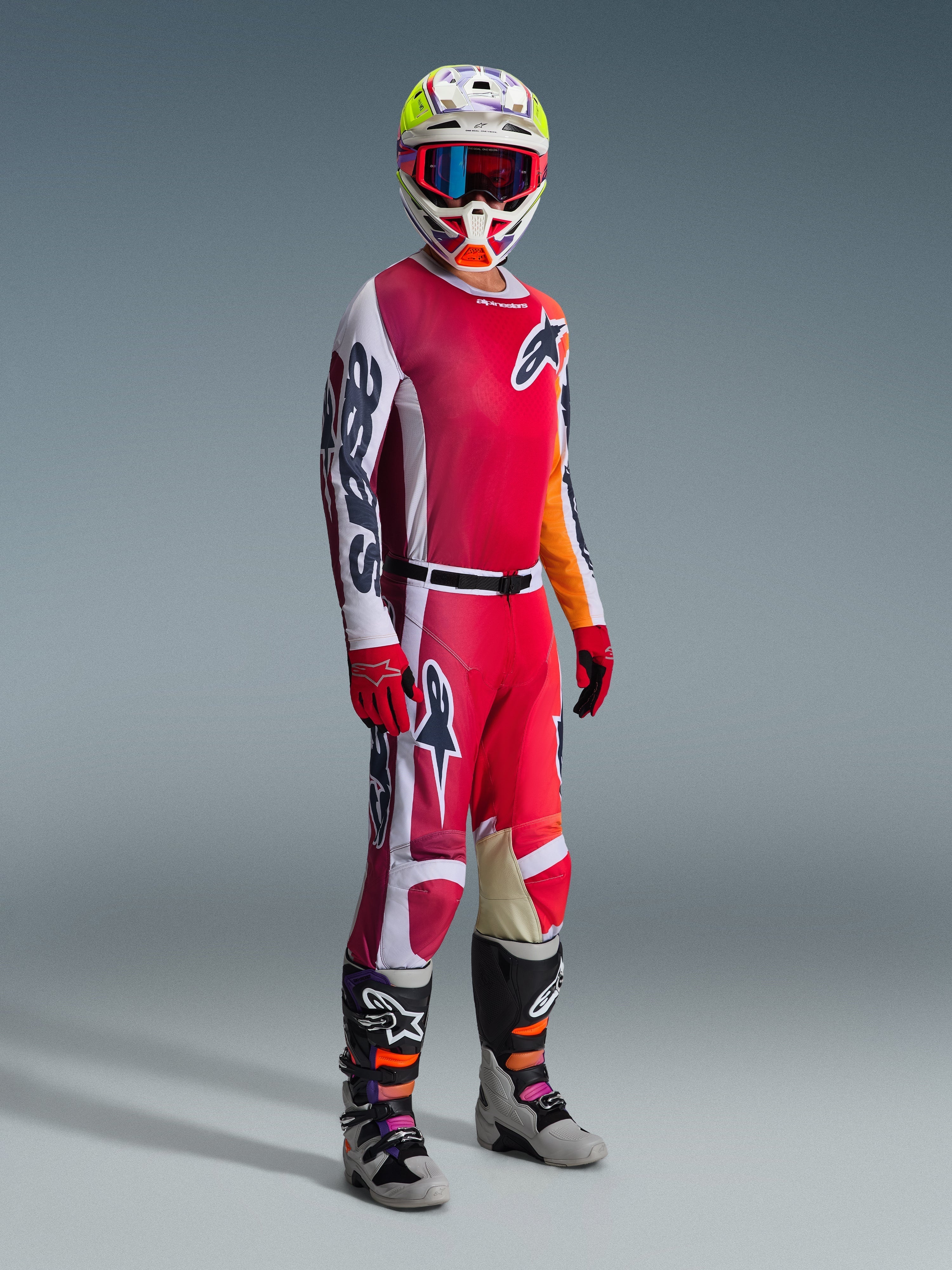 Pilota di motocross con abbigliamento completo da gara, maglia Alpinestars 2026 Racer Portl in rosso e grigio chiaro, pantaloni da motocross coordinati con accenti bianchi e verde scuro, casco multicolore con occhiali rossi, stivali grigi e neri con dettagli arancioni e viola, guanti rossi, outfit coordinato per corse off-road