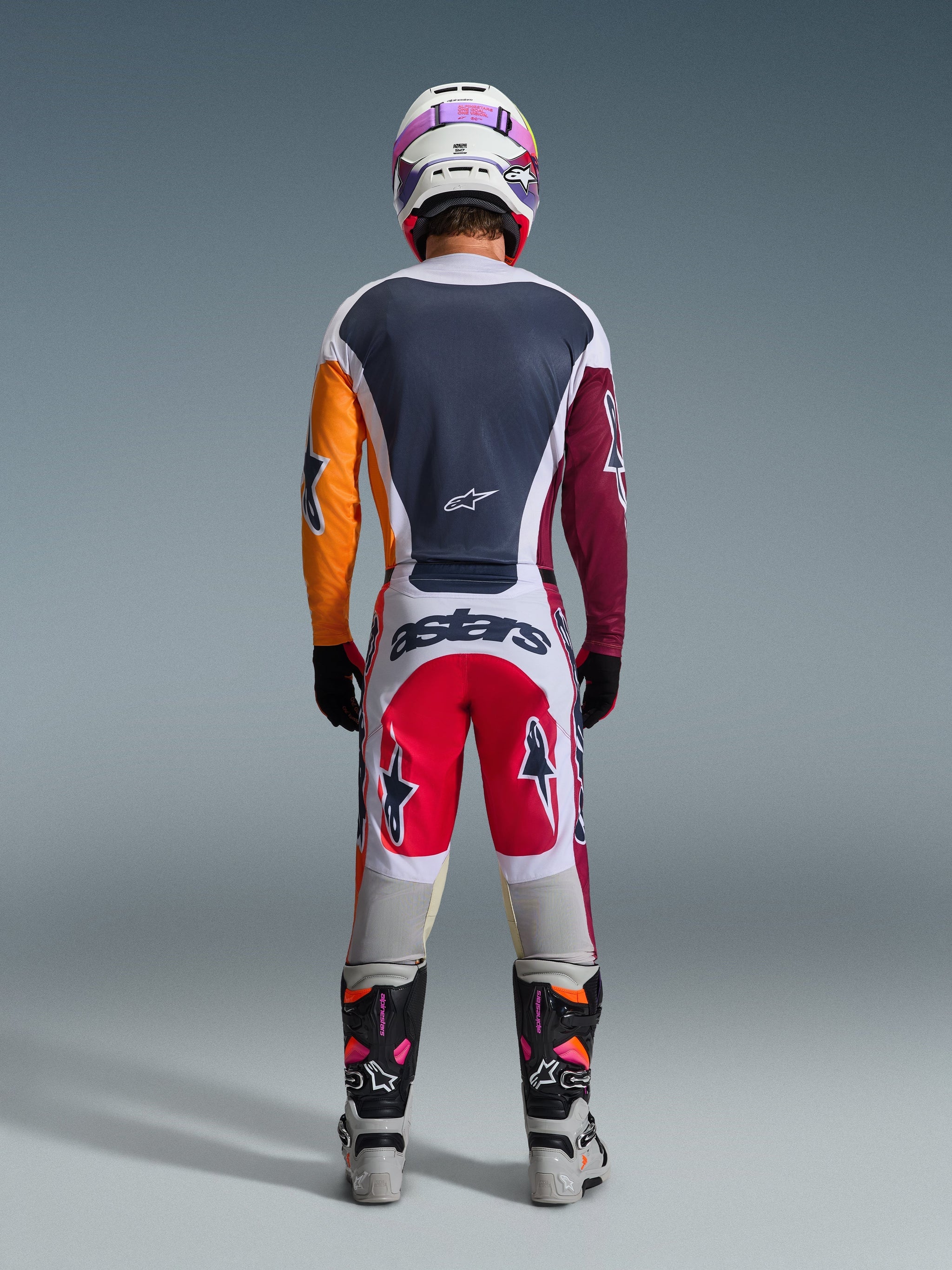 Pilota di motocross visto da dietro con abbigliamento completo da gara, Maglia Racer Portl 2026 in rosso, grigio chiaro e verde scuro con maniche asimmetriche arancione e bordeaux, pantaloni coordinati bianchi e rossi, casco bianco con maschera viola, stivali bianchi e neri, outfit coordinato per corse fuoristrada