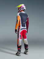 Pilota di motocross visto di spalle che indossa il completo Alpinestars Racer Portl 2026, caratterizzato da una maglia color-block con maniche arancioni e bordeaux, pantaloni bianchi con inserti rossi e branding 'astars' in evidenza, casco multicolore e stivali grigi, abbigliamento professionale da gara off-road.