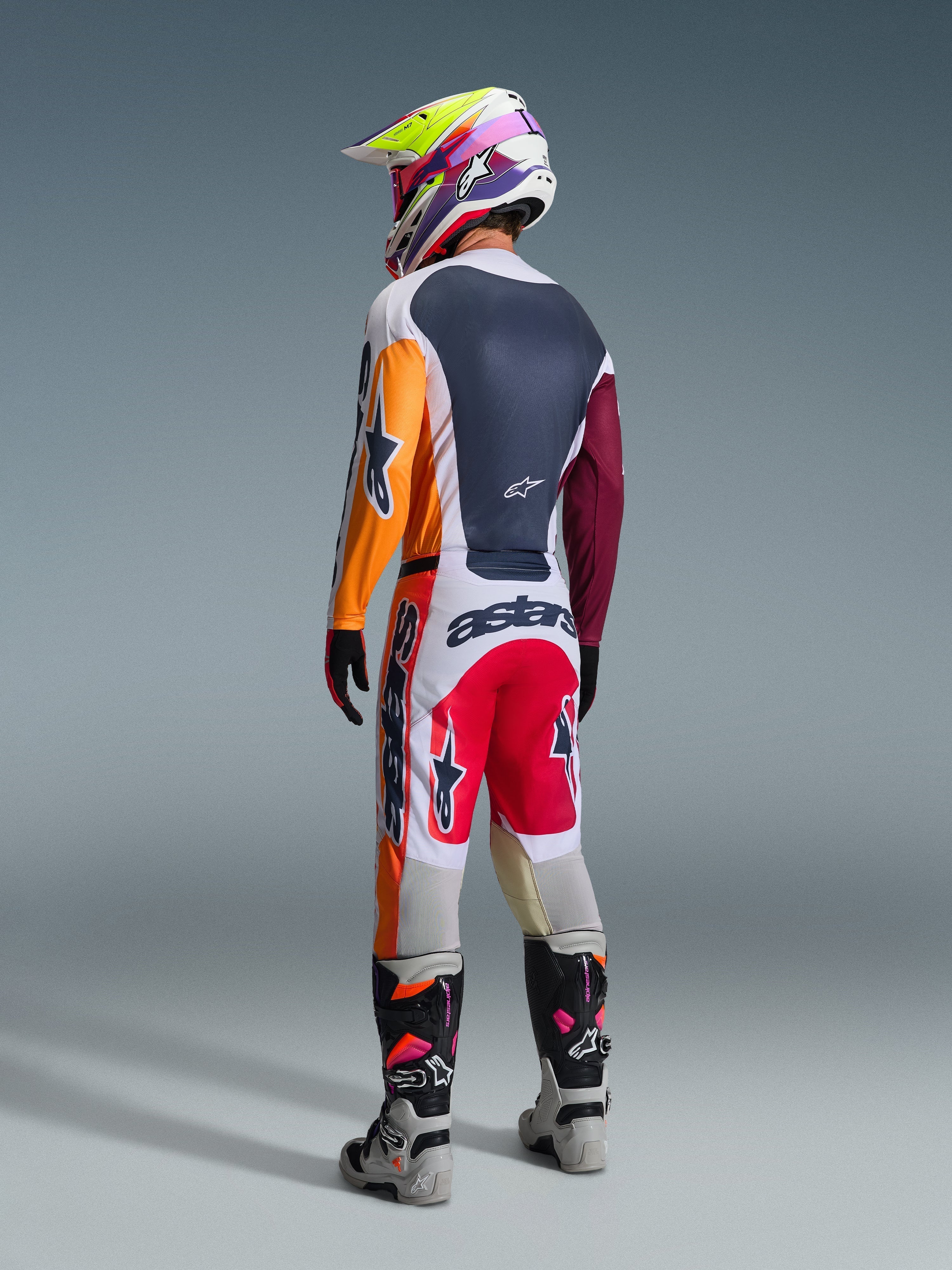 Pilota di motocross visto di spalle che indossa il completo Alpinestars Racer Portl 2026, caratterizzato da una maglia color-block con maniche arancioni e bordeaux, pantaloni bianchi con inserti rossi e branding 'astars' in evidenza, casco multicolore e stivali grigi, abbigliamento professionale da gara off-road.