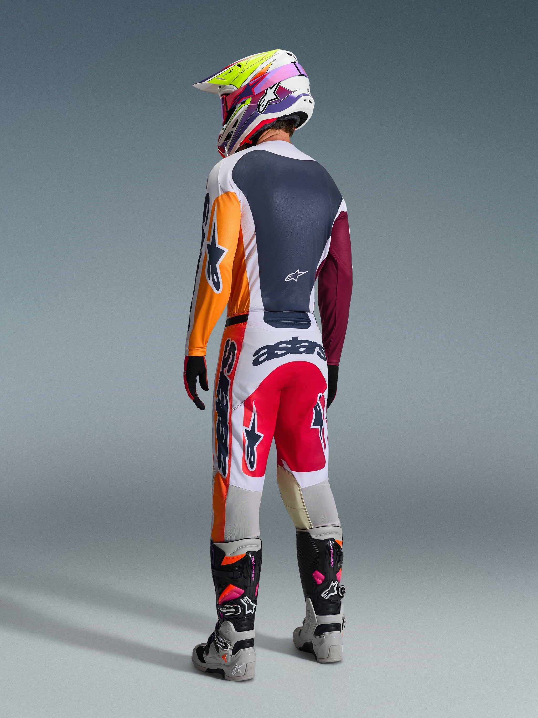 Pilota di motocross con abbigliamento da gara completo visto da dietro, Maglia Alpinestars 2026 Racer Portl in Rosso, Grigio Chiaro e Verde Scuro con pannelli arancioni e bordeaux, pantaloni bianchi e rossi coordinati, stivali grigi e neri con dettagli rosa, casco multicolore, completo da gara off-road coordinato.