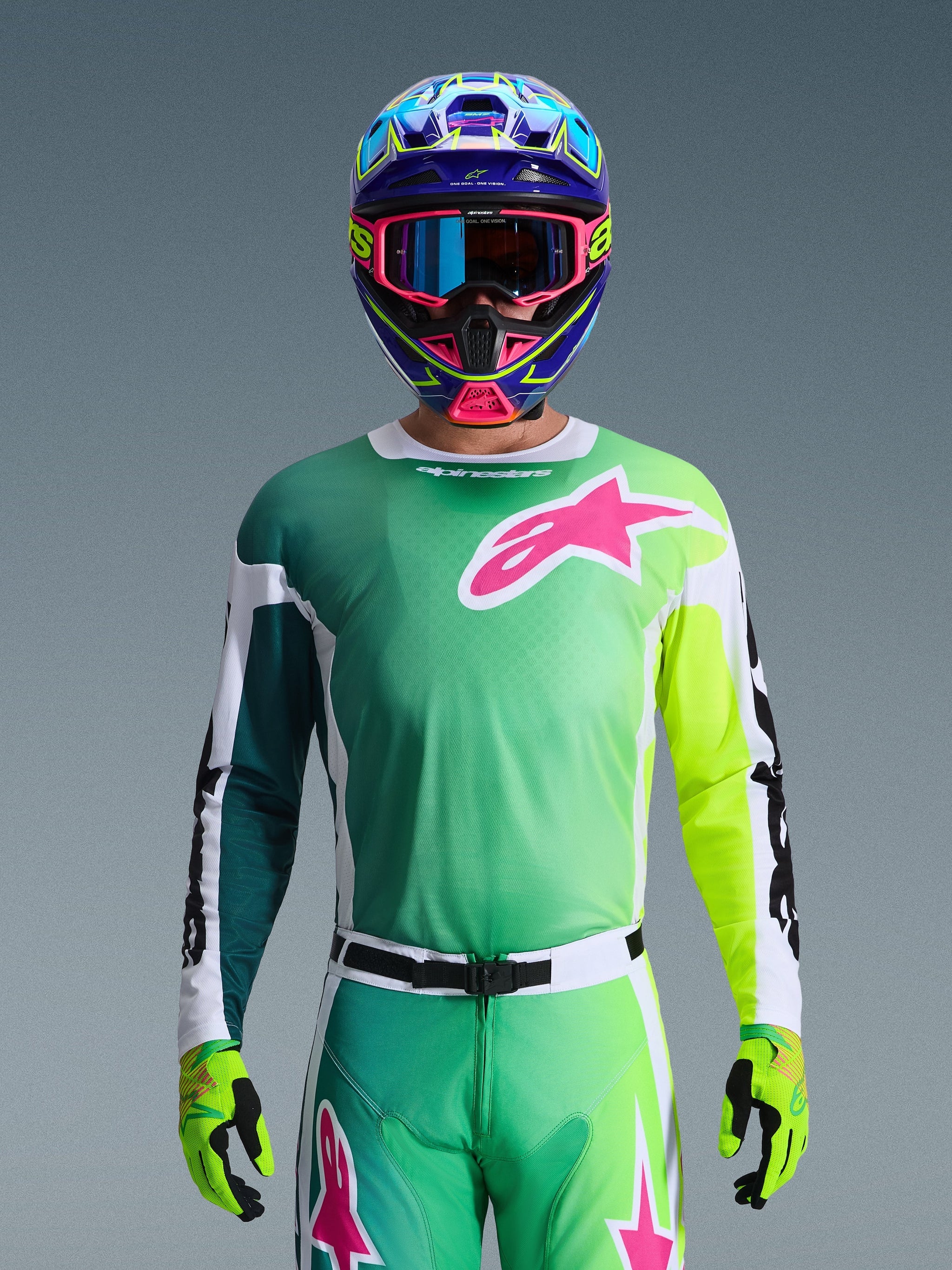 Alpinestars 2026 Racer Portl Jersey, maglia da motocross, verde, bianca e nera con un grande logo Alpinestars rosa sul petto, indossata da un pilota con un casco multicolore vivace e occhiali abbinati, abbigliamento professionale per corse fuoristrada