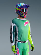 Maglia Alpinestars Racer Portl 2026, maglia da motocross, verde, bianco e nero, indossata da un pilota con pantaloni verdi coordinati, guanti fluorescenti e un casco blu e viola vivace con occhiali rosa, con logo Astars rosa sul petto.