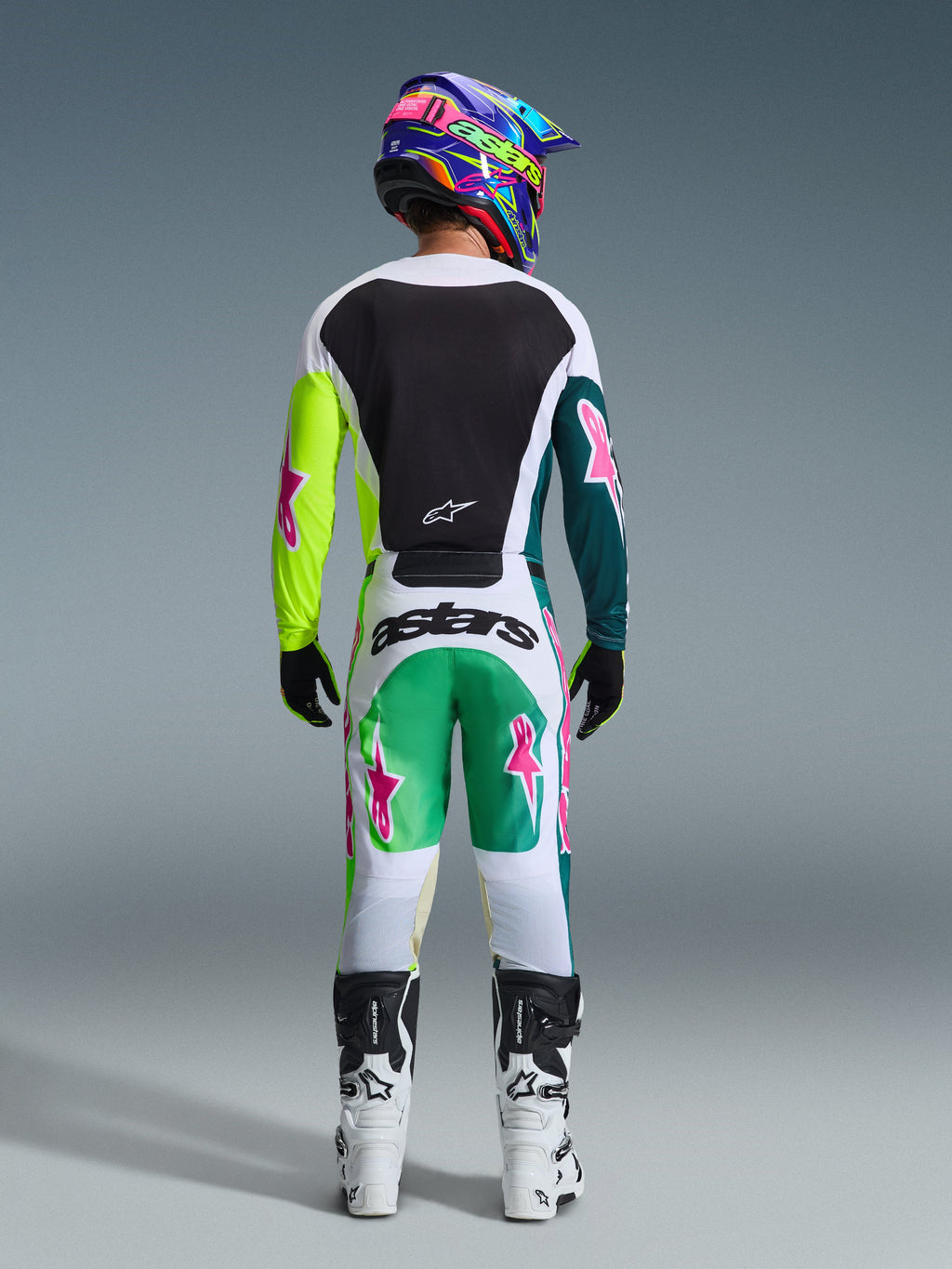 Vista posteriore di un pilota di motocross che indossa il completo Alpinestars 2026 Racer Portl, maglia bianca con pannello posteriore nero e maniche giallo neon e verde scuro con stelle rosa, pantaloni bianchi coordinati con dettagli verdi e teal e loghi a stella rosa, abbinati a un casco colorato, guanti neri e stivali bianchi e neri.