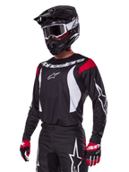 Pilota di motocross che indossa il set Alpinestars 2025 Fluid Haul, maglia nera con pannelli rossi sulle spalle e grafiche bianche, pantaloni neri coordinati con loghi a stella bianchi, guanti rossi e neri, casco nero con dettagli bianchi e occhiali, abbigliamento da motocross coordinato.