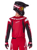 Maglia Alpinestars 2025 Fluid Haul, maglia da motocross, rosso acceso e nero, indossata come parte di un set completo con pantaloni e guanti coordinati, casco nero con occhiali a lente gialla, branding e loghi bianchi, abbigliamento da corsa fuoristrada ad alte prestazioni.