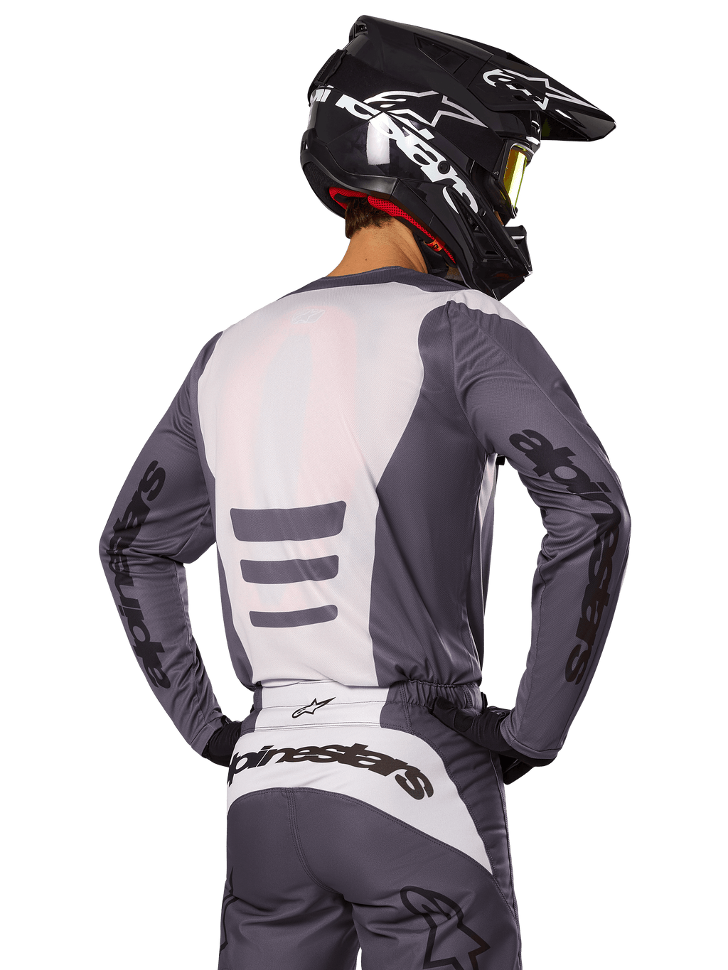 Pilota di motocross visto di spalle con set di abbigliamento Alpinestars Fluid Haul 2025, maglia e pantaloni grigi e bianchi, casco nero con grafiche bianche, maschera gialla, completo da gara coordinato