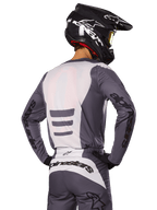 Pilota di motocross visto di spalle con set di abbigliamento Alpinestars Fluid Haul 2025, maglia e pantaloni grigi e bianchi, casco nero con grafiche bianche, maschera gialla, completo da gara coordinato
