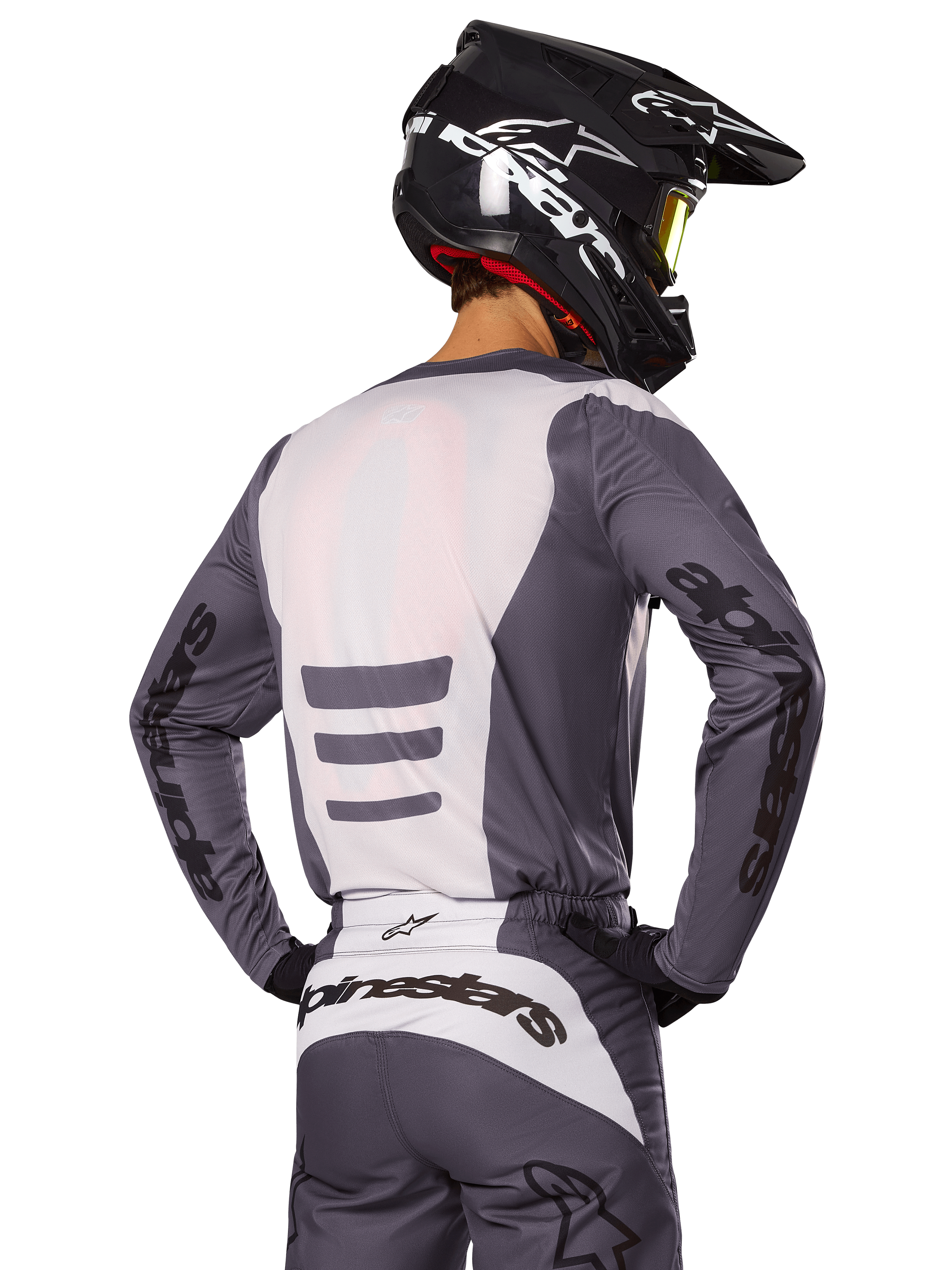 Pilota di motocross visto di spalle con set di abbigliamento Alpinestars Fluid Haul 2025, maglia e pantaloni grigi e bianchi, casco nero con grafiche bianche, maschera gialla, completo da gara coordinato