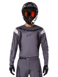 Pilota di motocross con maglia e set di abbigliamento Fluid Haul 2025, grigio con accenti neri e bianchi, maniche lunghe e loghi evidenti, casco nero con occhiali arancioni e guanti neri, outfit coordinato per corse fuoristrada