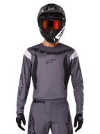 Pilota di motocross con maglia e set di abbigliamento Fluid Haul 2025, grigio con accenti neri e bianchi, maniche lunghe e loghi evidenti, casco nero con occhiali arancioni e guanti neri, outfit coordinato per corse fuoristrada