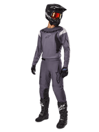 Pilota di motocross con set Alpinestars Fluid Haul 2025 grigio, maglia e pantaloni grigi con dettagli neri, casco nero con occhiali rossastri, guanti neri e stivali neri con loghi bianchi, completo da gara off-road