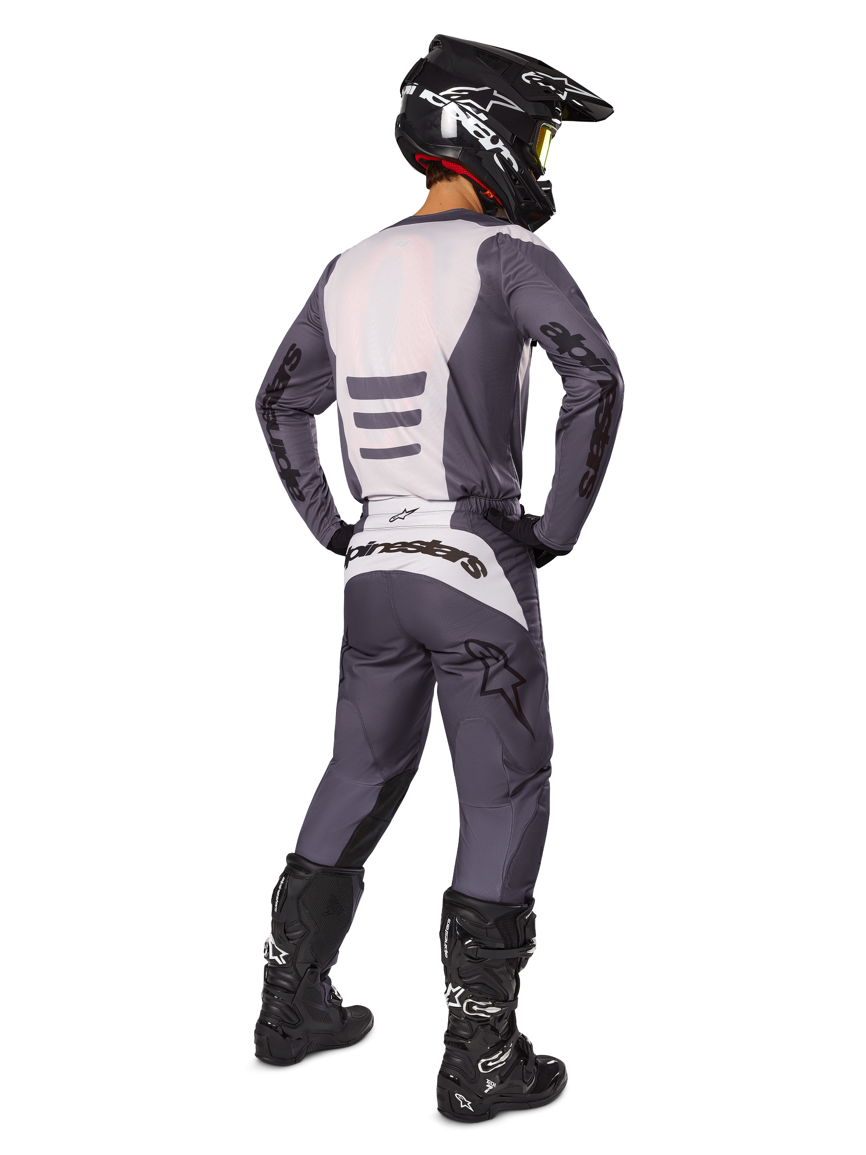 Pilota di motocross visto da dietro che indossa il set di abbigliamento Alpinestars 2025 Fluid Haul, maglia grigia e bianca e pantaloni coordinati, casco nero con occhiali gialli, stivali da motocross neri, equipaggiamento da gara completo per prestazioni fuoristrada