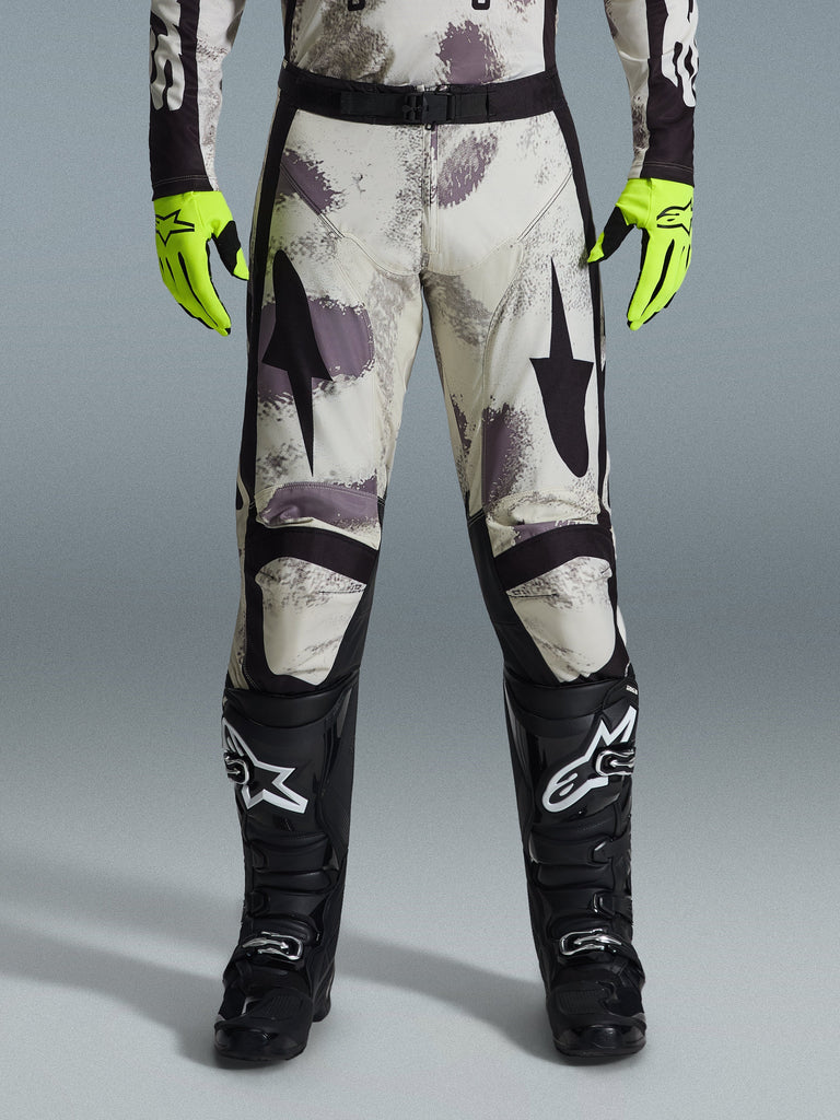 Alpinestars 2026 Racer Lahnd Pants, pantaloni da motocross, motivo Sand Camo con grafiche astratte grigie e viola, indossati con guanti giallo neon e stivali neri, con loghi a stella neri sulle cosce e cintura nera in vita, abbigliamento da gara off-road resistente