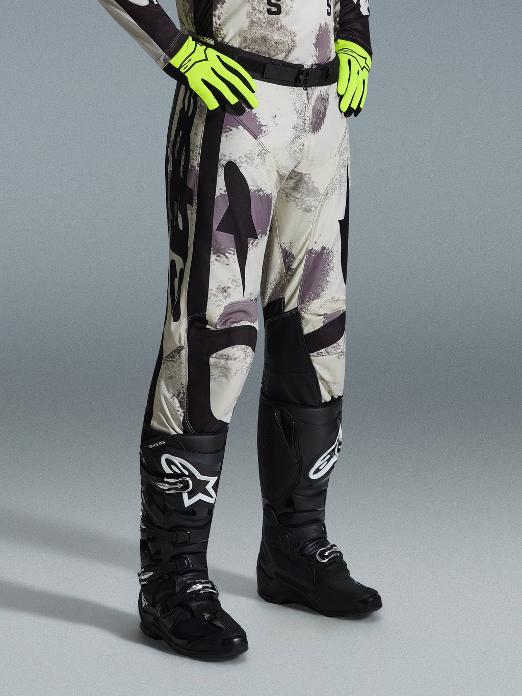 Alpinestars 2026 Racer Lahnd Pants, pantaloni da motocross, Sand Camo, indossati con guanti giallo neon e stivali da motocross neri, caratterizzati da un motivo camouflage astratto con pannelli laterali neri, design ergonomico off-road