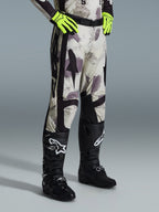 Alpinestars 2026 Racer Lahnd Pants, pantaloni da motocross, Sand Camo, indossati con guanti giallo neon e stivali da motocross neri, caratterizzati da un motivo camouflage astratto con pannelli laterali neri, design ergonomico off-road