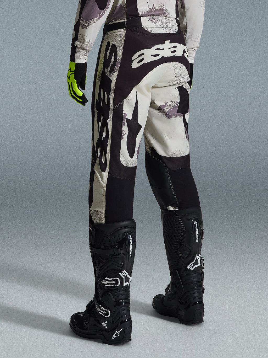 Alpinestars 2026 Racer Lahnd Pants, pantaloni da motocross, Sand Camo e grigio, vista posteriore con marchio bianco evidente sulla seduta e sulle gambe, abbinati a stivali da motocross neri e guanti gialli, abbigliamento professionale per fuoristrada