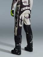 Alpinestars 2026 Racer Lahnd Pants, pantaloni da motocross, Sand Camo e grigio, vista posteriore con marchio bianco evidente sulla seduta e sulle gambe, abbinati a stivali da motocross neri e guanti gialli, abbigliamento professionale per fuoristrada